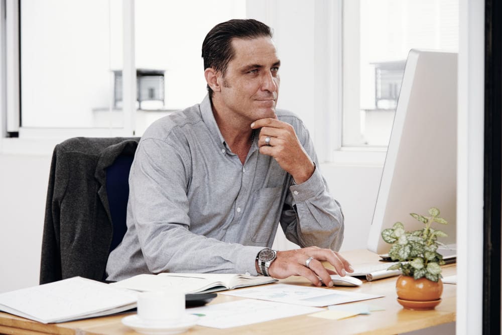Homem sentado em frente ao computador, pensando com a mão no queixo
