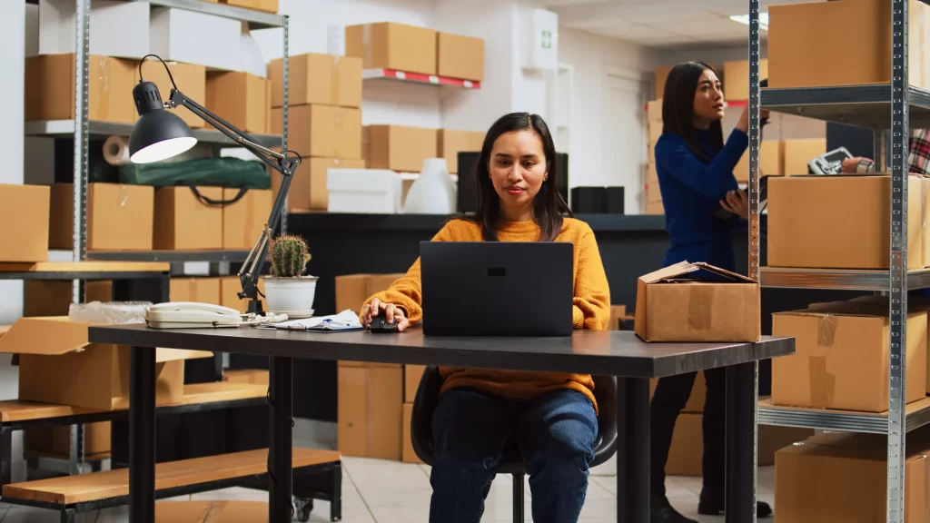 Vendas em e-commerce com duas mulheres, uma usando notebook e outra no estoque com caixas, representando gestão online e logística