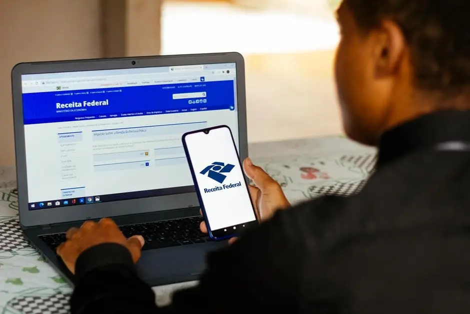 Rapaz consultando pendências no site da Receita Federal pelo notebook e aplicativo no celular.